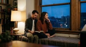 A man reads a book beside a woman gazing thoughtfully out a rainy window in a cozy apartment at dusk